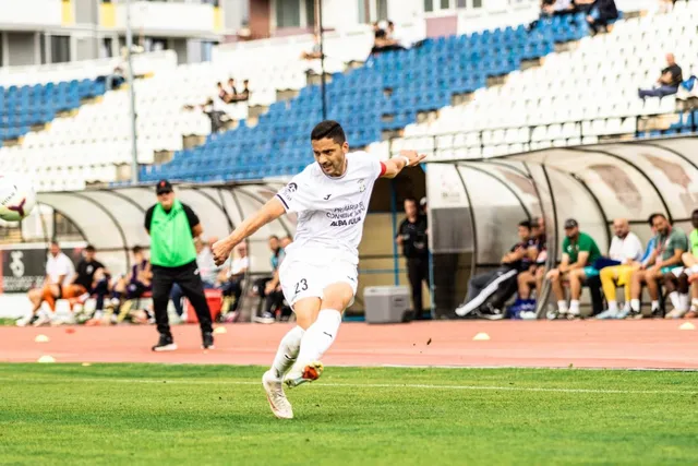 Un jucător de fotbal îmbrăcat în alb lovește mingea pe un teren de iarbă verde în fața unor tribune cu scaune albastre și albe.