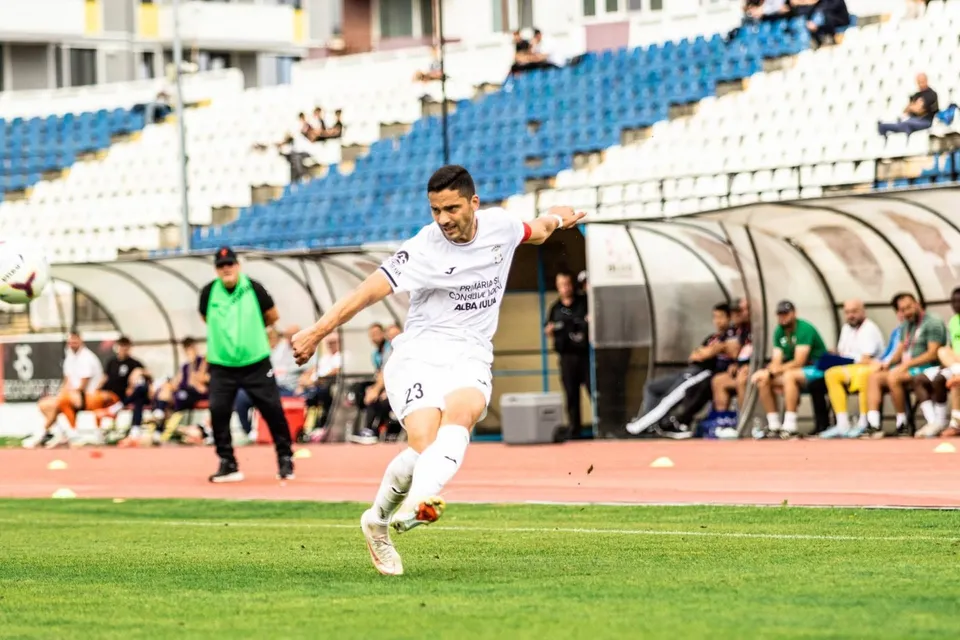 Un jucător de fotbal îmbrăcat în alb lovește un balon pe un teren de iarbă verde în fața unor tribune cu scaune albastre.