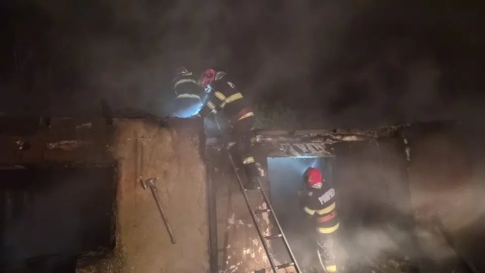 Trei pompieri cu echipament de protecție și cască sunt poziționați pe o scară și lângă o clădire înconjurată de fum noaptea.