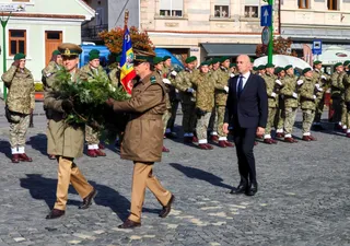 A group of military personnel in uniform and a man in a dark suit walk on a paved street during a formal ceremony, with one officer carrying a large green wreath.