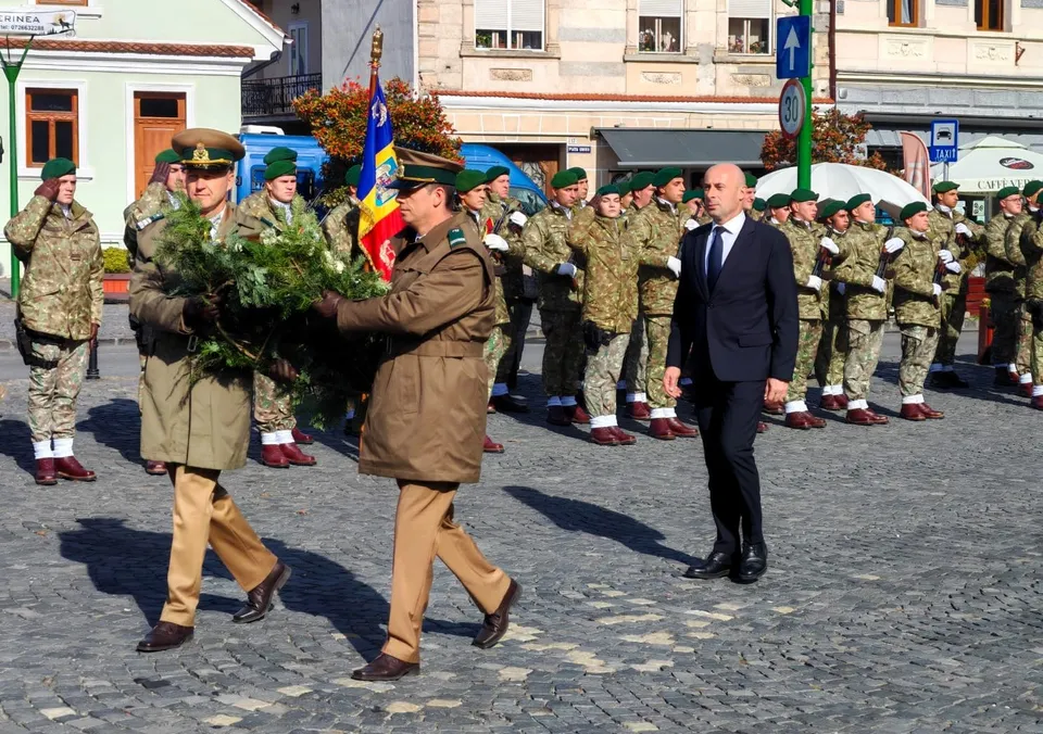 A group of military personnel in uniform and a man in a dark suit walk on a paved street during a formal ceremony, with one officer carrying a large green wreath.