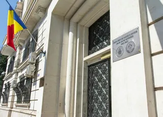 O clădire albă cu ferestre protejate de gratii metalice negre, având un steag tricolor și o placă metalică pe perete.
