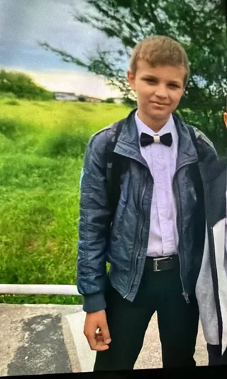Un băiat cu părul scurt poartă o cămașă albă, papion negru, o jachetă albastră și pantaloni negri stând în fața unui câmp verde.