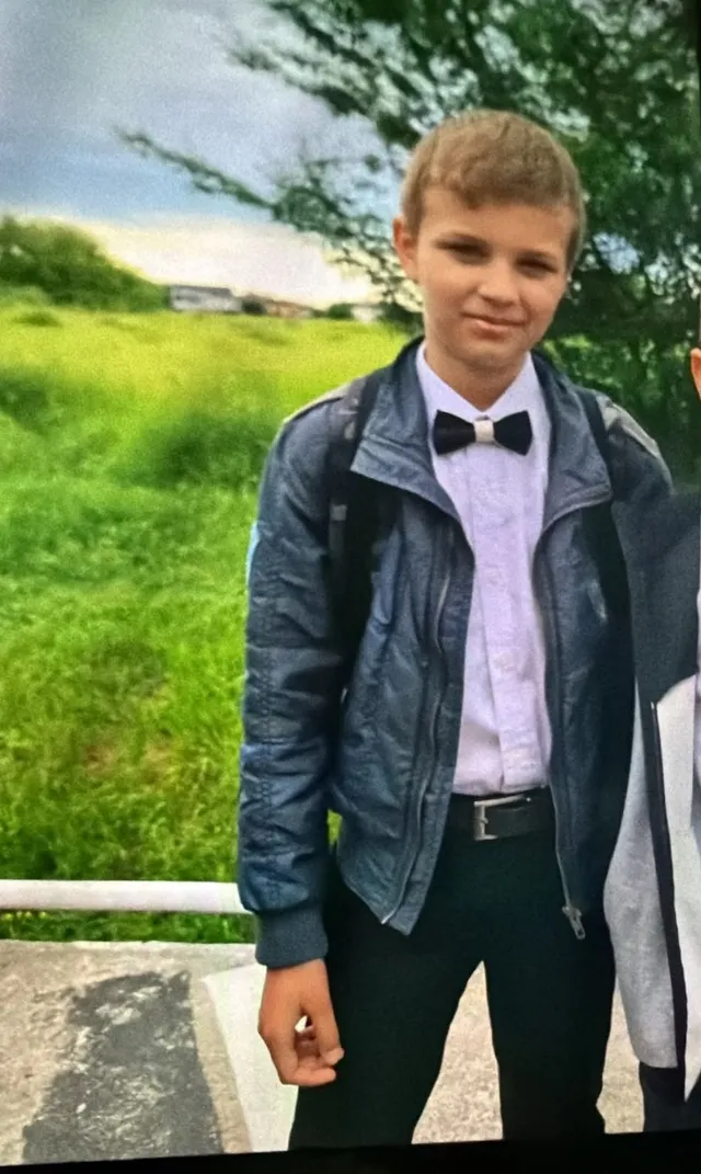 Un băiat cu părul scurt poartă o cămașă albă, papion negru, o jachetă albastră și pantaloni negri stând în fața unui câmp verde.