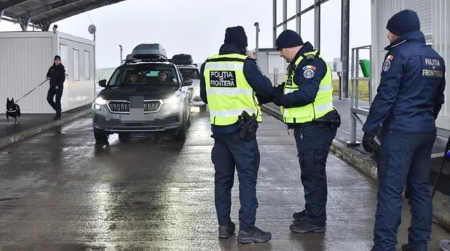 Trei polițiști în uniformă cu marcaje de frontieră stau lângă o mașină gri pe o suprafață asfaltată umedă, în timp ce un câine și un om sunt vizibili.