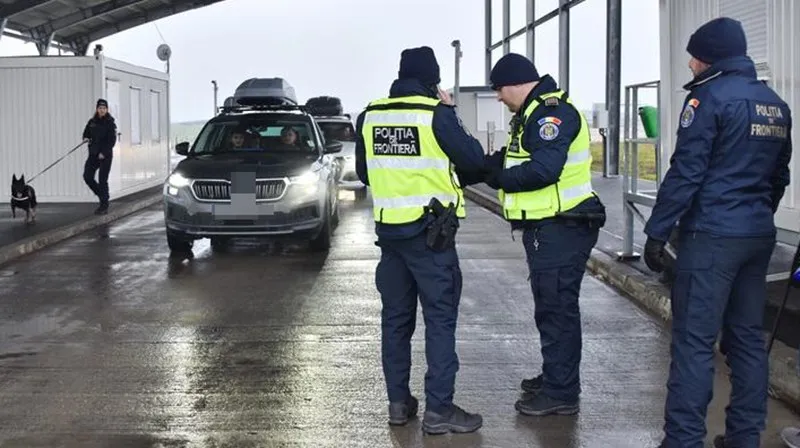 Trei polițiști în uniformă cu marcaje de frontieră stau lângă o mașină gri pe o suprafață asfaltată umedă, în timp ce un câine și un om sunt vizibili.