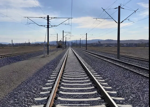 Două șine de tren cu traverse de beton sunt dispuse pe un balast de pietriș sub linii electrice aeriene și un cer înnorat.