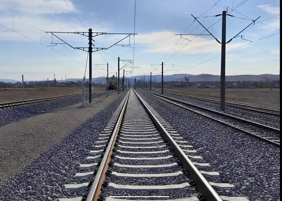 Două șine de tren cu traverse de beton sunt dispuse pe un balast de pietriș sub linii electrice aeriene și un cer înnorat.