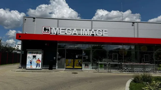 O clădire de magazin cu un panou roșu și inscripția MEGA IMAGE este situată sub un cer cu nori albi.