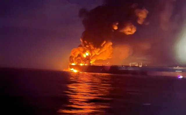 Un vas mare arde pe apă sub un cer întunecat, generând flăcări portocalii și un nor dens de fum negru.