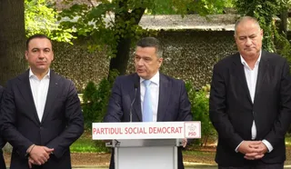 Trei bărbați în costume purtând cămăși albe stau în fața unui podium alb pe care scrie Partidul Social Democrat.