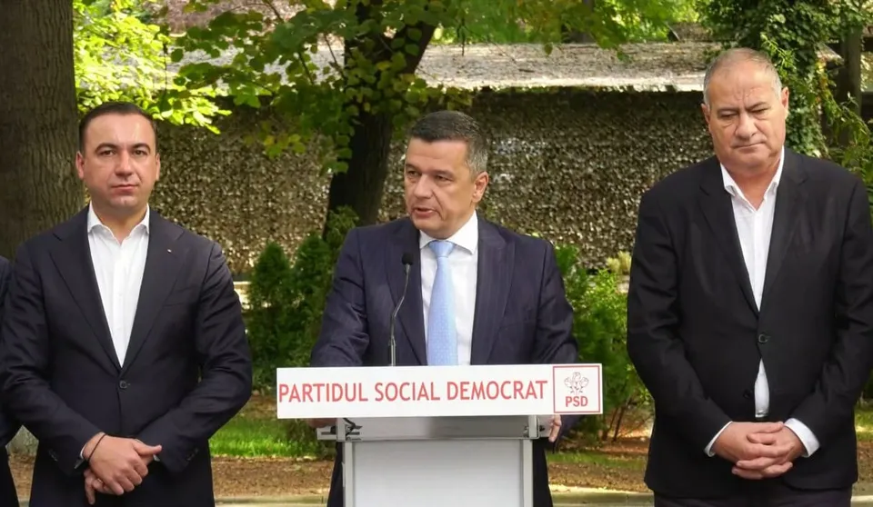 Trei bărbați în costume purtând cămăși albe stau în fața unui podium alb pe care scrie Partidul Social Democrat.