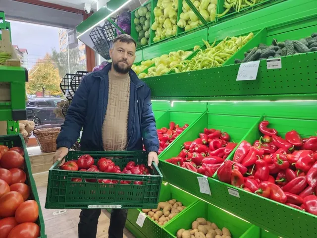 Un bărbat cu barbă poartă un coș verde cu legume roșii în fața unor rafturi verzi pline cu diverse fructe și legume.