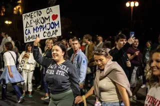 O mulțime de oameni merge pe un drum noaptea, iar o femeie ridică un panou alb cu inscripții negre și un simbol de inimă roșie.