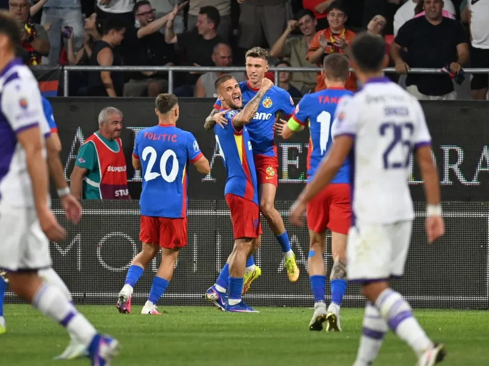 Câțiva fotbaliști în echipamente albastre și roșii sărbătoresc pe un teren de fotbal în fața unor spectatori din tribune.