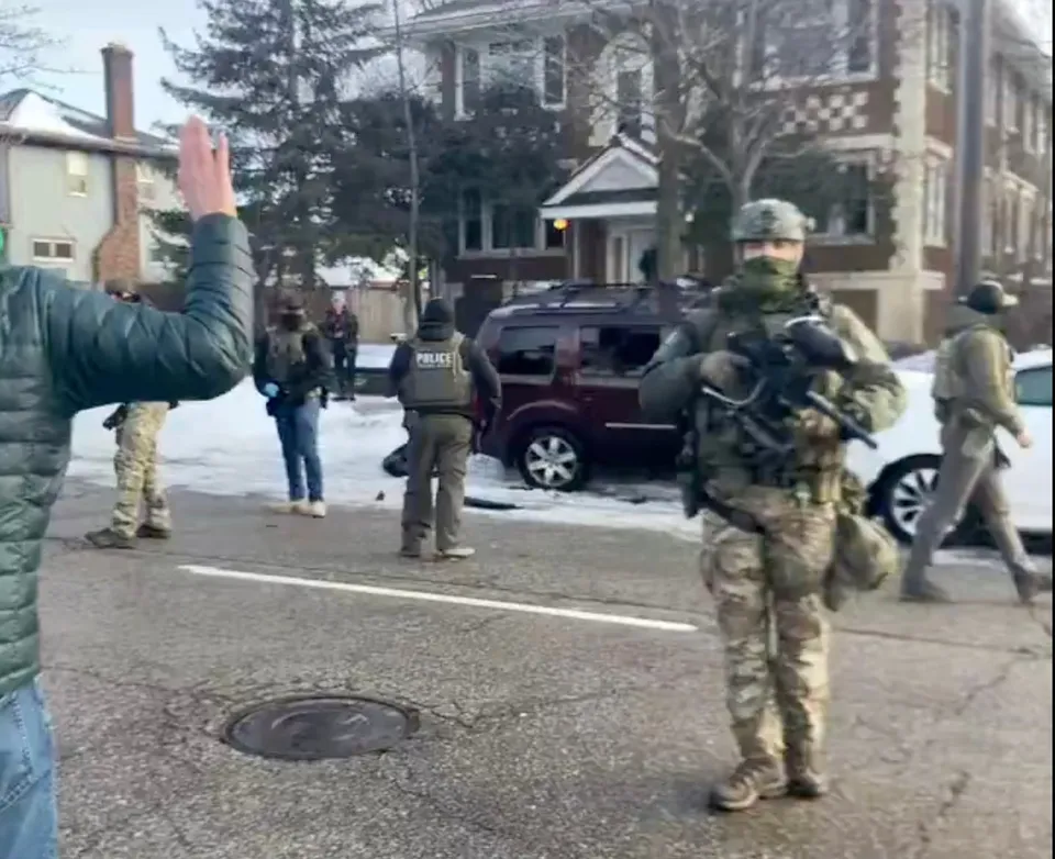 Mai mulți bărbați în uniforme militare și de poliție stau pe o stradă acoperită parțial de zăpadă lângă un vehicul de culoare închisă.