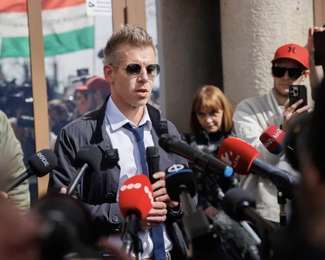 Un bărbat cu ochelari de soare și cravată albastră stă în fața mai multor microfoane de jurnalism într-un spațiu exterior.