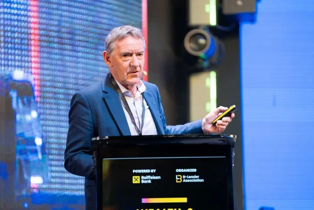 Un bărbat cu sacou albastru și cămașă albă stă în spatele unui podium negru, ținând un dispozitiv mobil în mână.