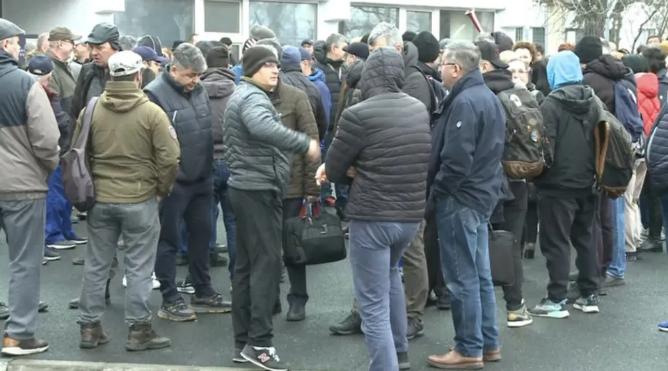 Un grup de persoane îmbrăcate în haine de iarnă stă adunat pe o stradă lângă o clădire cu ferestre mari.