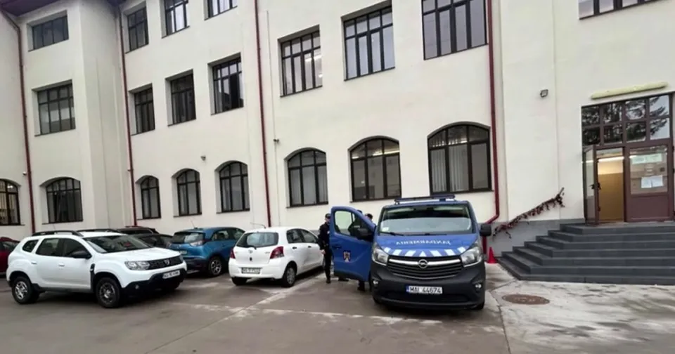 Mai multe mașini sunt parcate în fața unei clădiri albe cu ferestre mari, lângă scări și o intrare cu uși maronii.