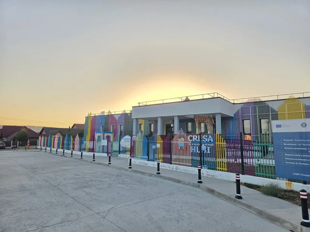 O clădire colorată cu un gard metalic în față este situată lângă o stradă asfaltată sub un cer luminos la apus.