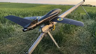 O dronă neagră cu anvergură mare și insigne albastre-galbene stă pe un suport metalic într-un câmp verde.