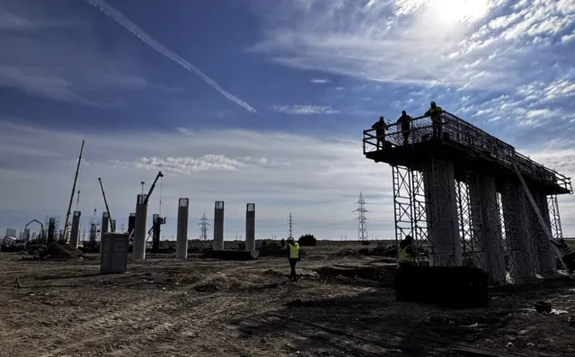 Siluete ale unor muncitori stau pe o platformă înaltă de construcție lângă piloni de beton sub un cer înzăpezit de nori.