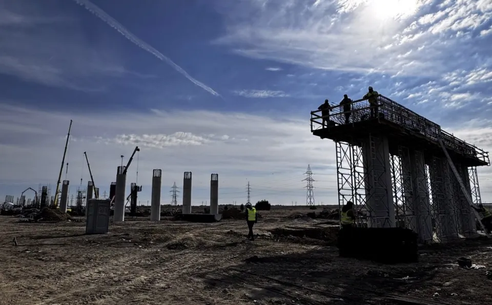 Siluete ale unor muncitori stau pe o platformă înaltă de construcție lângă piloni de beton sub un cer înzăpezit de nori.