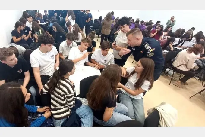 Un grup de tineri stă așezat pe scaune și pe podea într-o încăpere, în timp ce un bărbat în uniformă întinde mâna spre unul dintre ei.