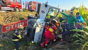 Echipaje de salvatori în uniforme colorate lucrează lângă o autoutilitară albă răsturnată într-un câmp de porumb lângă un camion de pompieri roșu.