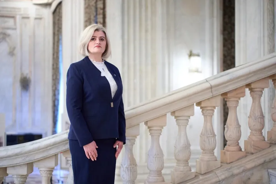 O femeie cu părul blond, purtând un costum albastru închis, stă pe o scară de marmură cu balustradă sculptată într-un interior iluminat.