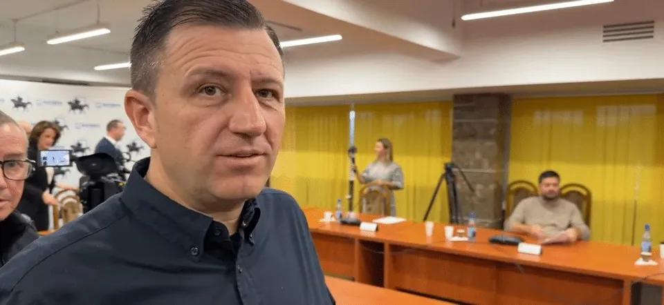 Un bărbat cu cămașă închisă la culoare privește spre cameră, în timp ce în fundal se văd persoane la mese într-o încăpere cu perdele galbene.