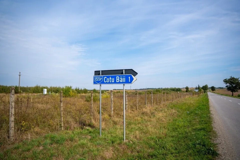 Un semn de circulație albastru cu textul Cotu Băii indică direcția pe lângă un drum asfaltat și un câmp cu iarbă uscată sub un cer senin.