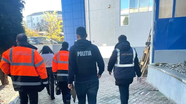 Cinci persoane purtând haine de lucru portocalii și jachete închise cu inscripții merg pe un trotuar lângă o clădire cu panouri albastre.
