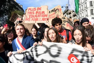 Un grup de tineri strigă și ține diverse pancarte din carton și un banner alb în timpul unei manifestări stradale sub cerul senin.