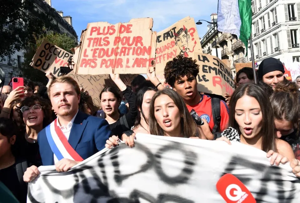 Un grup de tineri strigă și ține diverse pancarte din carton și un banner alb în timpul unei manifestări stradale sub cerul senin.