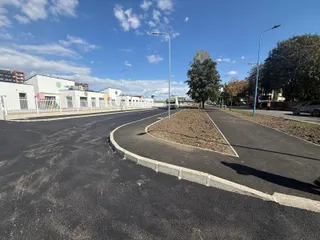 O porțiune de asfalt proaspăt turnat cu borduri de beton se întinde spre un teren cu pământ și clădiri albe sub un cer cu nori.