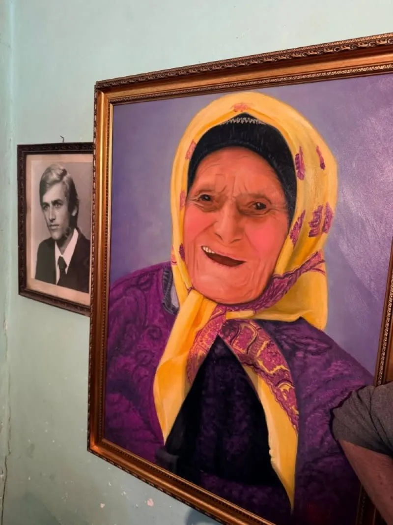 Un tablou încadrat în lemn prezintă portretul unei femei cu un basma galbenă și o rochie mov pe un fundal violet.
