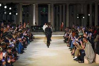 O persoană merge pe o pistă de catwalk în fața unui public care folosește telefoane mobile pentru fotografiat.