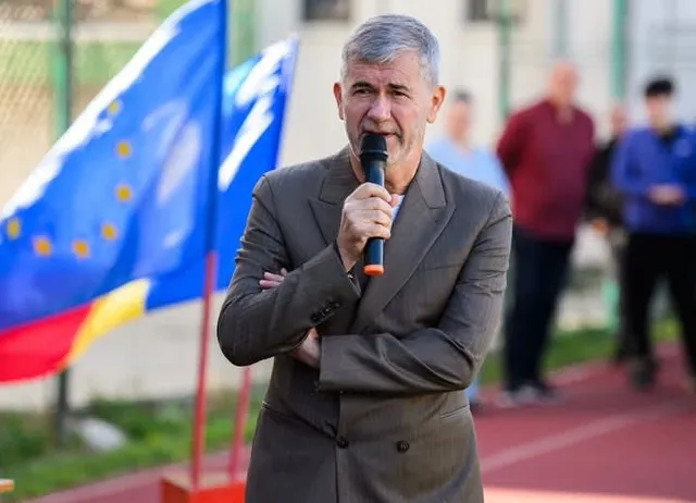 Un bărbat cu părul cărunt, îmbrăcat într-un sacou maro, ține un microfon negru cu vârful portocaliu în fața feței în timp ce vorbește.