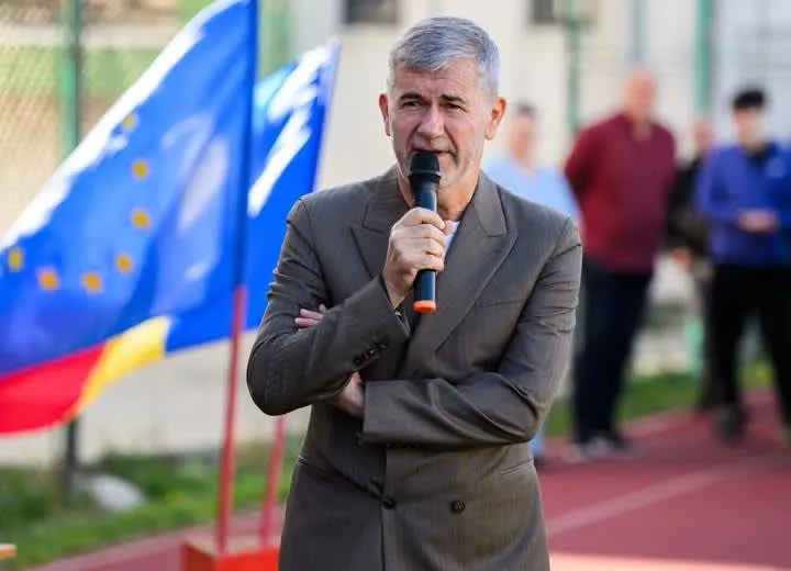 Un bărbat cu părul cărunt, îmbrăcat într-un sacou maro, ține un microfon negru cu vârful portocaliu în fața feței în timp ce vorbește.