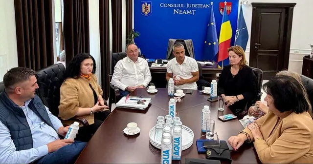 O grupă de persoane stă la o masă de conferințe de formă dreptunghiulară, pe care se află sticle de apă, căni și documente.