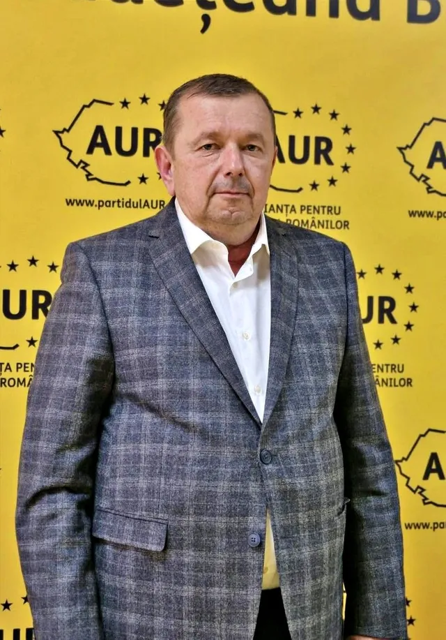Un bărbat cu părul scurt și scurtă barbă poartă un sacou gri cu model și o cămașă albă în fața unui fundal galben cu logouri AUR.