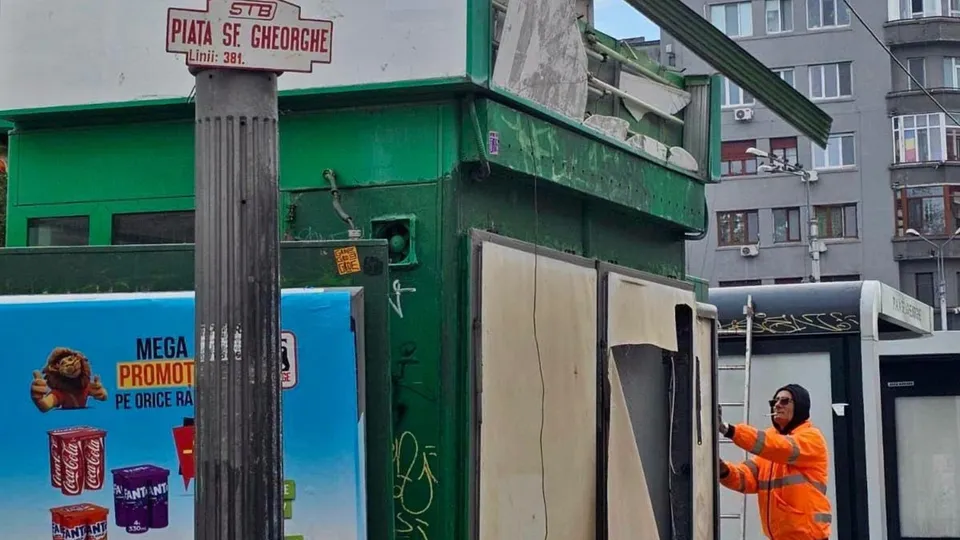 O persoană în vestimentație portocalie cu benzi reflectorizante stă lângă o structură verde cu panouri de lemn și un stâlp cu un indicator.