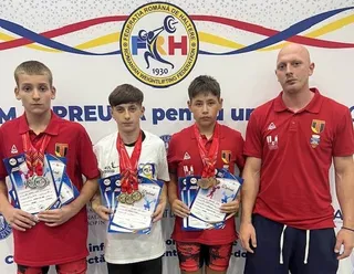 Trei băieți cu medalii și diplome stau în linie lângă un bărbat îmbrăcat într-un tricou roșu în fața unui panou cu un logo circular.