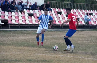 Un jucător în echipament albastru și alb conduce mingea pe un teren de fotbal în fața unui adversar îmbrăcat în roșu și albastru.