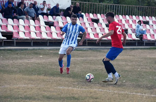 Un jucător în echipament albastru și alb conduce mingea pe un teren de fotbal în fața unui adversar îmbrăcat în roșu și albastru.