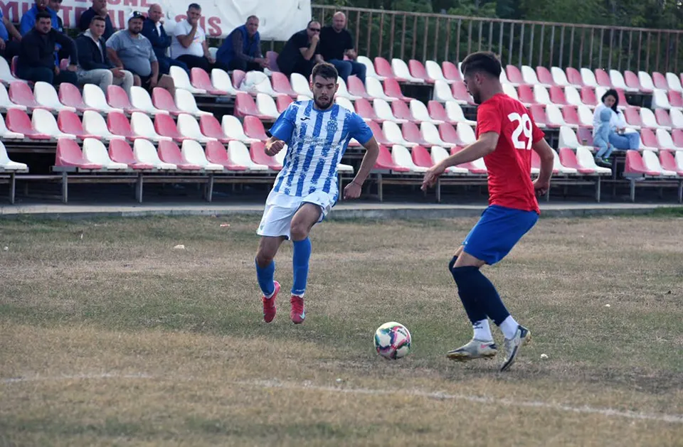 Un jucător în echipament albastru și alb conduce mingea pe un teren de fotbal în fața unui adversar îmbrăcat în roșu și albastru.