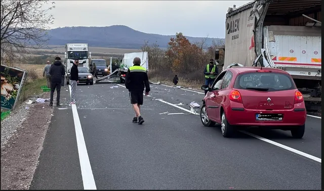 Un vehicul roșu și un camion mare stau pe o șosea asfaltată lângă persoane care se află în apropierea unor vehicule de marfă.