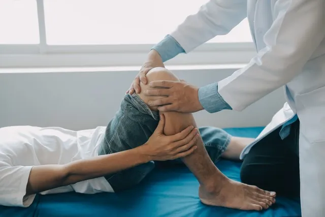 Un medic într-o halat albă examinează genunchiul unui pacient care stă întins pe o masă de examinare albastră.